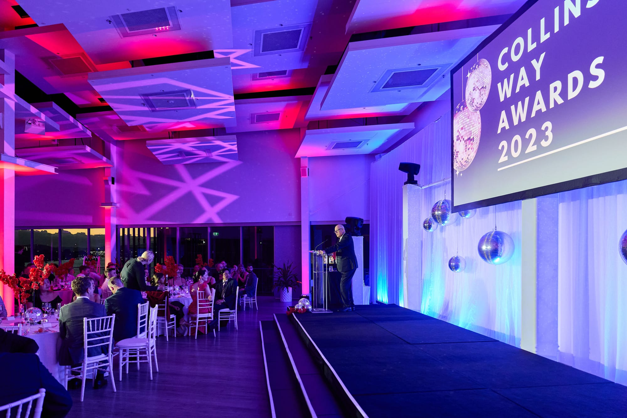 A large event space set for an awards dinner, with banquet rounds, a stage and a projection screen