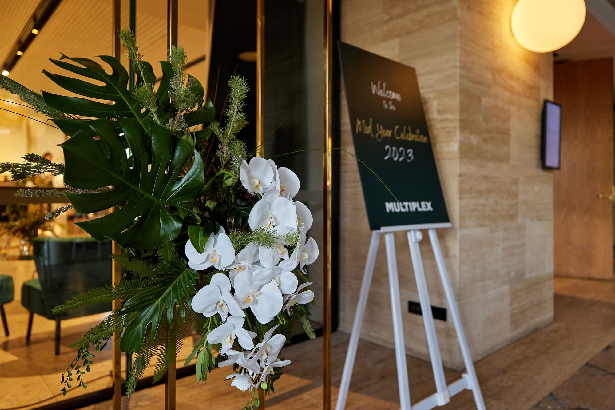 The entrance to a hotel function room: a welcome sign on an easel naming a company mid-year celebration, brass-framed glass doors, a white orchid and monstera arrangement in the foreground and a warm globe light on the stone wall