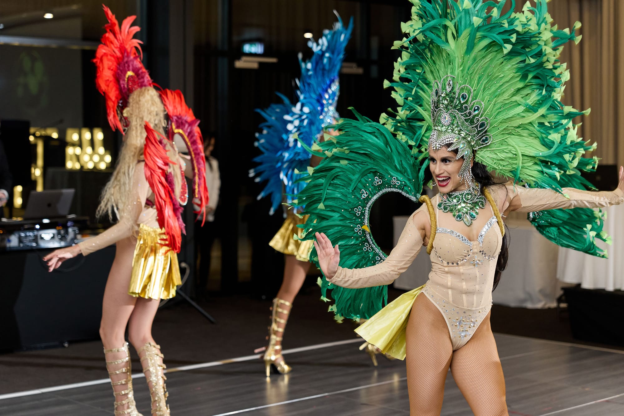 Three samba dancers in red, blue and green feathered headdresses perform on the dance floor at an evening function, with a DJ desk behind them