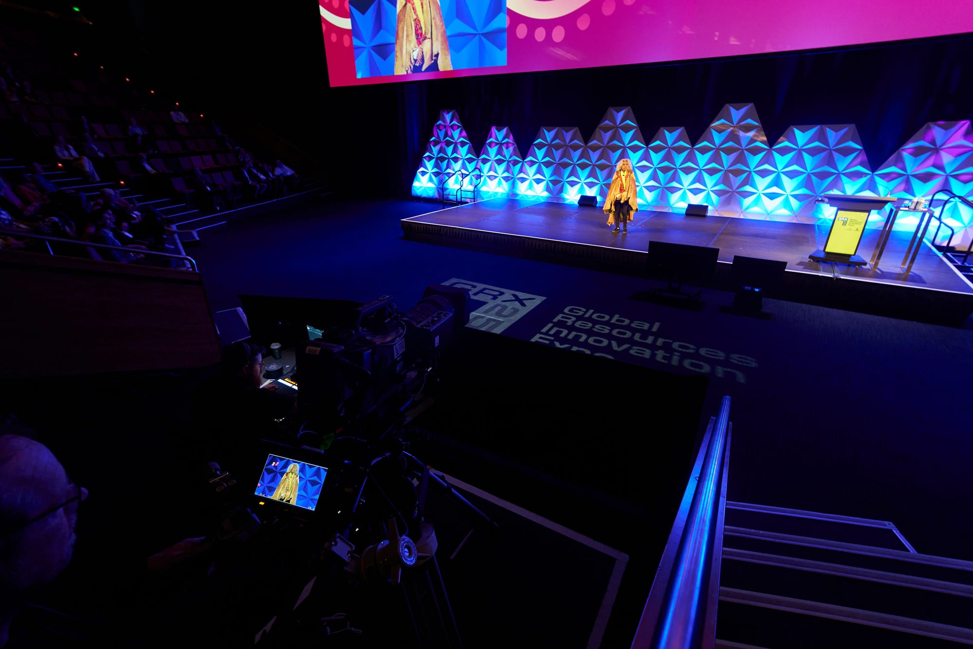 A camera operator on a riser films a speaker on a lit conference stage, with a large projection screen above the stage and the audience in raked seating to the left