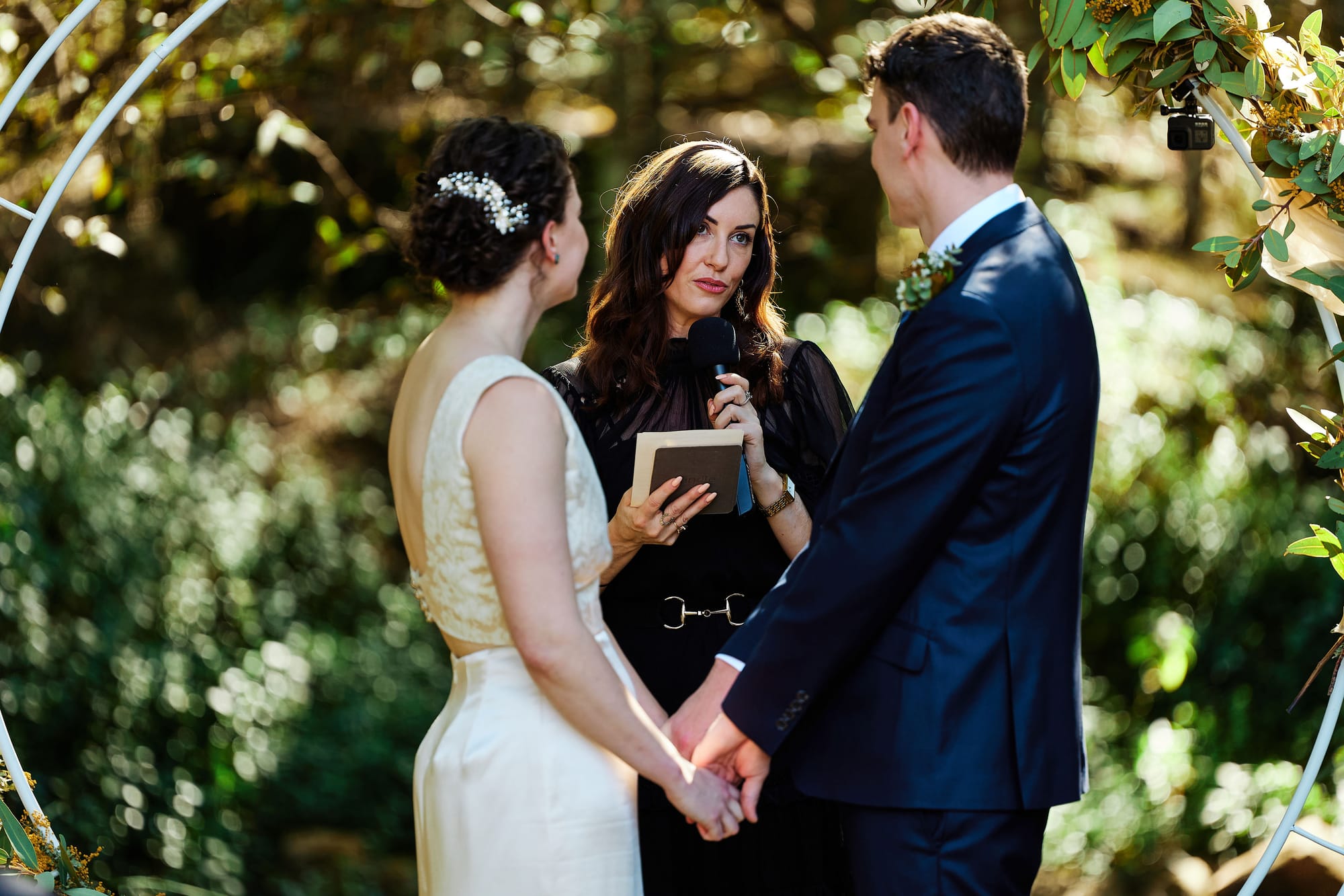 A celebrant holding a microphone and her ceremony notes speaks to a couple holding hands under a round floral arbor in a sunlit garden