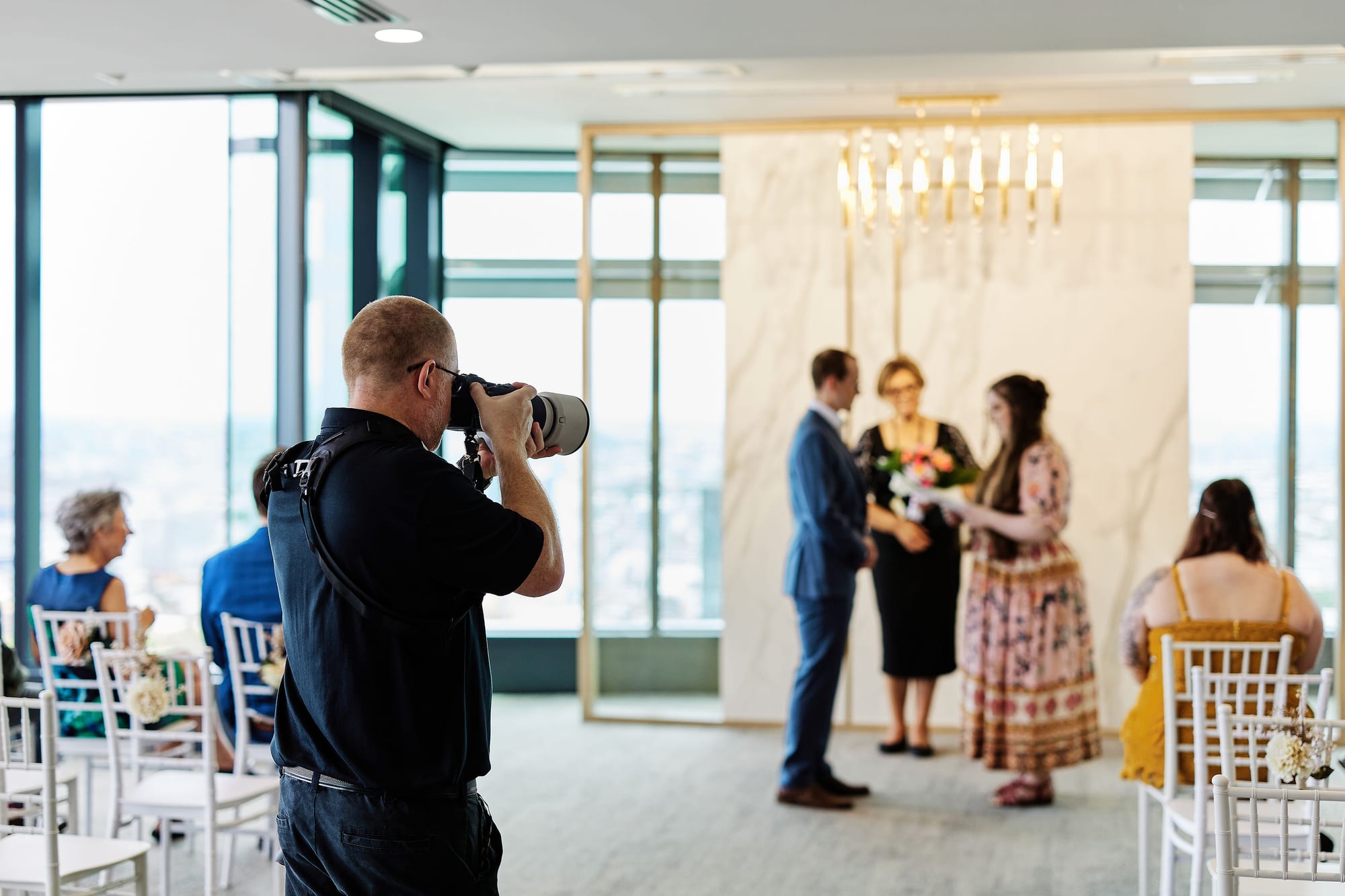 A photographer in the foreground shoots a wedding ceremony in a high-rise function room, the couple and celebrant standing before a marble wall and gold chandelier while guests watch from white chairs